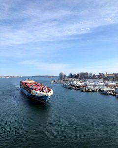 Photograph of a ship carrying shipping containers into port under blue slightly cloudy sky.